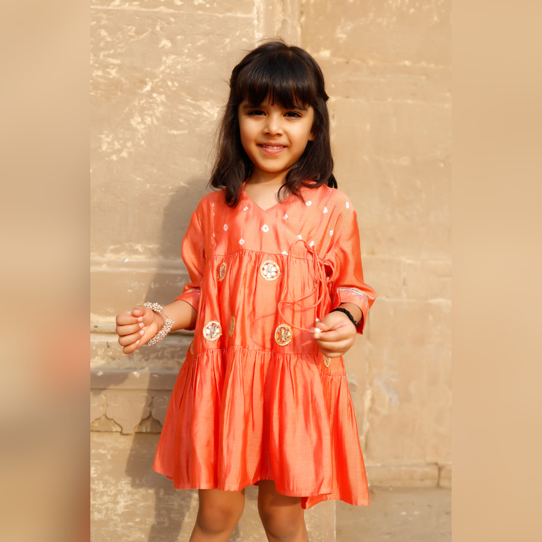 Little girl in a vibrant orange ethnic dress adorned with laces and gota flowers.